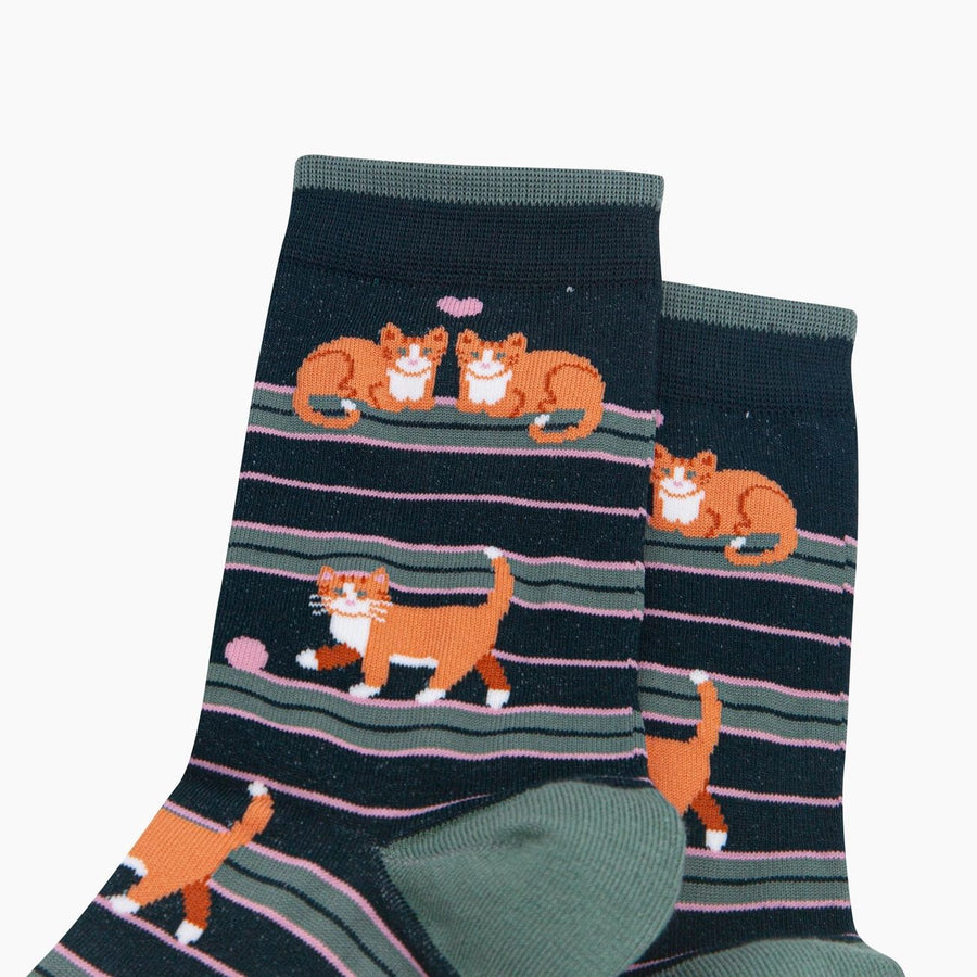 A pair of dark navy crew socks with a pattern of orange and white cats walking on pink and gray horizontal stripes, shown on a plain white background.