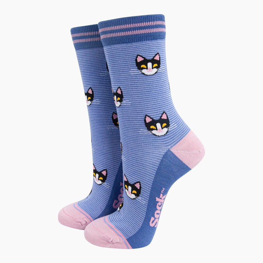 A pair of crew socks in a medium blue striped pattern, featuring a repeating pattern of black and white cat faces with pink inner ears and yellow eyes.