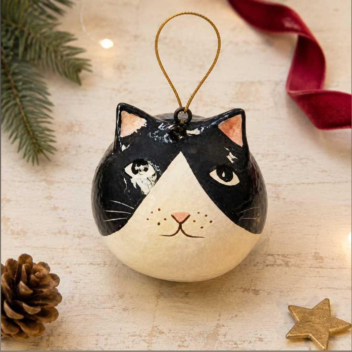 A hand-painted cat-shaped Christmas ornament with a black and cream face, hanging by a gold string, styled on a light wooden surface with pine branches