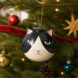 A hand-painted Christmas ornament shaped like a black and white cat's face, hanging from a gold string on a decorated Christmas tree with red ribbon and lights.