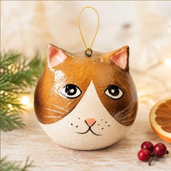 A cat-shaped Christmas ornament with a brown and cream face, black eyes, and a gold hanging loop, styled on a wooden surface with holiday greenery