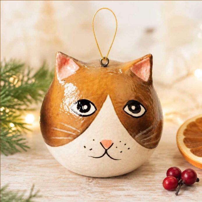 A cat-shaped Christmas ornament with a brown and cream face, black eyes, and a gold hanging loop, styled on a wooden surface with holiday greenery