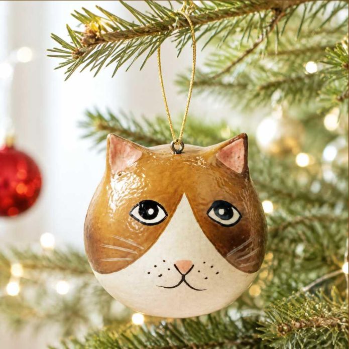 A hand-painted cat-shaped Christmas ornament hanging from a pine tree branch with warm bokeh lights in the background.