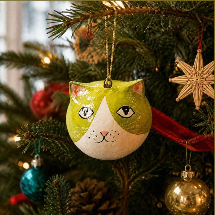 A hand-painted Christmas ornament shaped like a cat's head hangs on a decorated Christmas tree. The ornament has a green and white painted design with simple black eyes and a pink nose