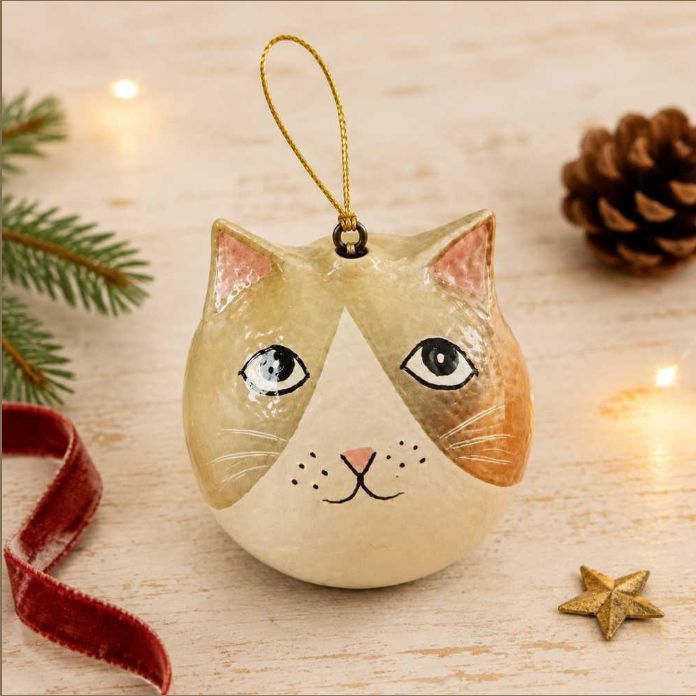 A cat-shaped Christmas ornament with a gold hanging loop, decorated with beige, cream, and orange-brown tones, set on a light wooden surface with festive props