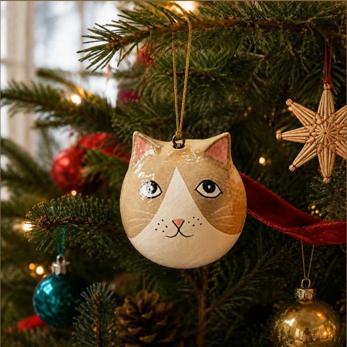 A round cat-face Christmas ornament hanging from a pine tree branch, surrounded by other holiday decorations including a woven star, red ribbon, and colourful ornaments.