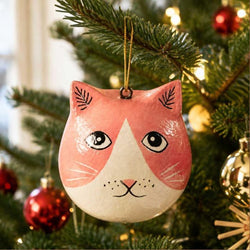 A hand-painted cat-shaped Christmas ornament hanging on a green Christmas tree with warm string lights and red ornaments in the background.