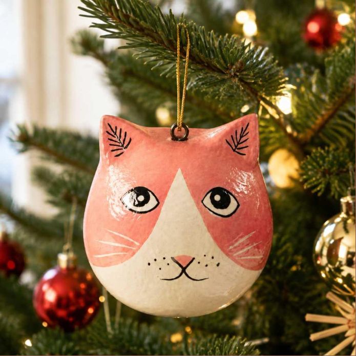 A hand-painted cat-shaped Christmas ornament hanging on a green Christmas tree with warm string lights and red ornaments in the background.