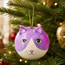 A round hand-painted Christmas ornament shaped like a cat's face, featuring purple and white coloring with black and gold details, hanging from a gold string
