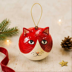 A glossy red and cream cat-shaped Christmas ornament with a gold hanging loop, styled on a light wooden surface with a red ribbon, pine branch, pinecone, and small gold star.