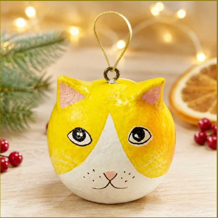 A round, hand-painted cat-shaped Christmas ornament with a yellow and white face, hanging by a gold string on a wooden surface with festive holiday decorations in the background.