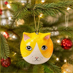 A hand-painted cat-shaped Christmas ornament hanging from a green Christmas tree branch, with warm holiday lights and red ornaments blurred in the background.