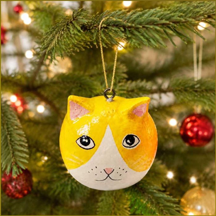 A hand-painted cat-shaped Christmas ornament hanging from a green Christmas tree branch, with warm holiday lights and red ornaments blurred in the background.