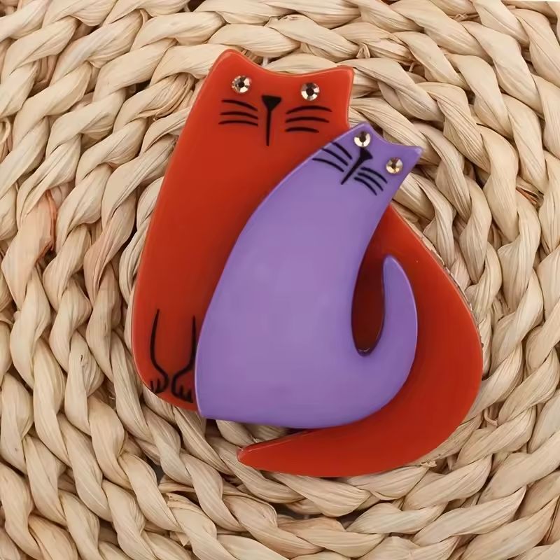 A two cat-shaped brooch, one red and one purple, on a woven surface