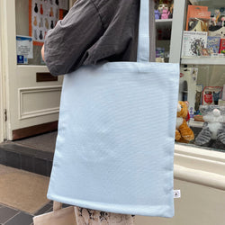 A person carries a large, plain light blue canvas tote bag with a matching shoulder strap in front of a shop window.