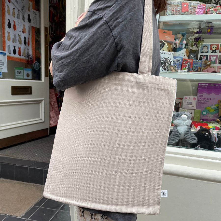 A person carries a large, plain beige canvas tote bag over their shoulder in front of a shop window.