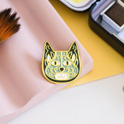 An enamel pin badge depicting a stylised cat face with a floral pattern in gold, black and white colours