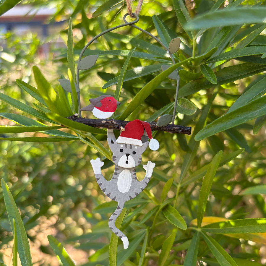 A small hanging Christmas ornament shaped like a grey cat wearing a red Santa hat, suspended from a twig branch