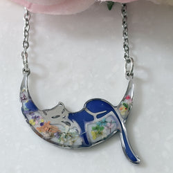 A crescent moon pendant necklace featuring a sleeping cat design with blue enamel, silver metal, and embedded dried flowers