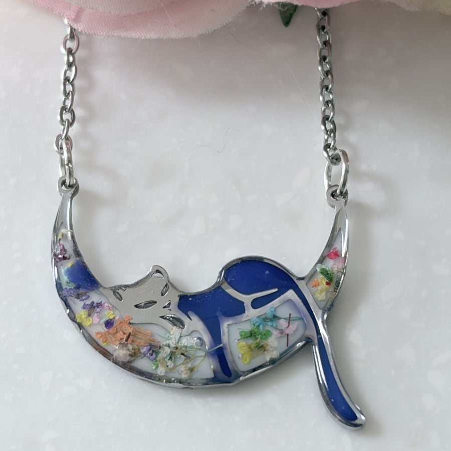 A crescent moon pendant necklace featuring a sleeping cat design with blue enamel, silver metal, and embedded dried flowers