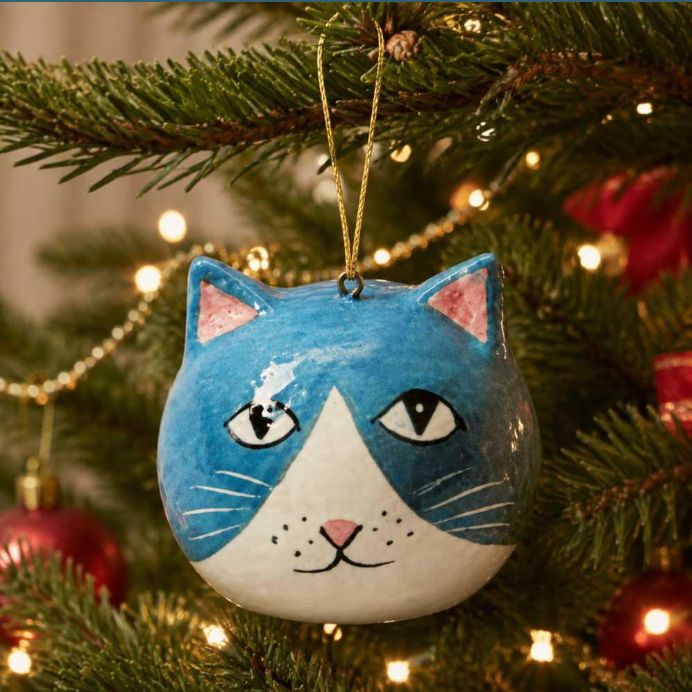 A round cat-shaped Christmas ornament with a blue and white face hangs on a decorated Christmas tree branch with warm string lights.