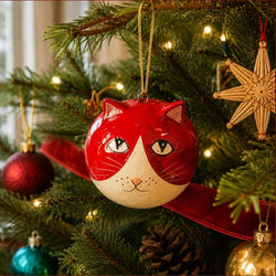 A round Christmas ornament shaped like a cat's head, painted red and cream, hanging on a decorated Christmas tree with warm lights and other ornaments.