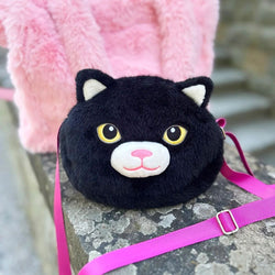 A round black plush cat face bag with yellow eyes, a white muzzle, and a pink nose, resting on a stone surface with a pink strap