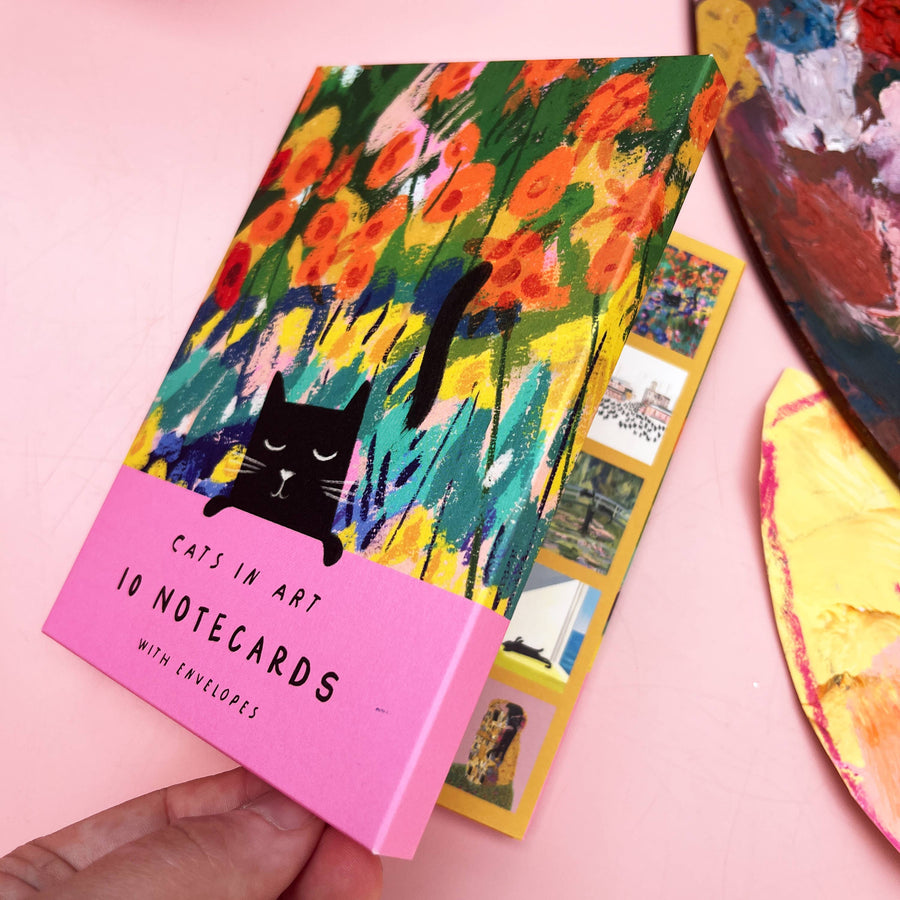 A hand holds a pink notebook card set titled "Cats in Art: 10 Notecards with Envelopes"