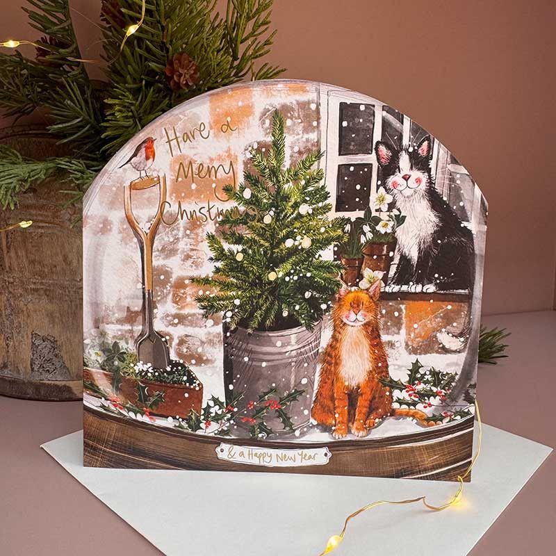 A festive Christmas greeting card with an arched top, featuring an illustrated winter scene with a potted Christmas tree, an orange cat, a black-and-white cat by a window, a shovel, and the text 'Have a Merry Christmas & a happy new year'. 