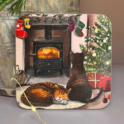 A square decorative coaster, printed with a cosy Christmas fireplace scene featuring two cats, a lit wood stove, stockings, a tree, and wrapped gifts