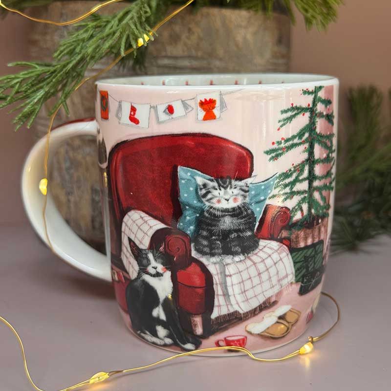 A ceramic mug with a festive Christmas illustration of cats on a red armchair, surrounded by soft pine branches and warm fairy lights.