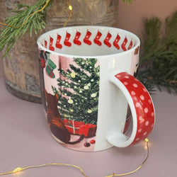 A white ceramic Christmas-themed mug with a red polka-dot handle and a festive print of a decorated Christmas tree, stockings, and a cat.