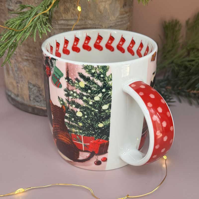 A white ceramic Christmas-themed mug with a red polka-dot handle and a festive print of a decorated Christmas tree, stockings, and a cat.