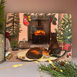A rectangular decorative placemat featuring a cozy illustrated winter scene with two cats, a fireplace, Christmas stockings, trees, and wrapped gifts