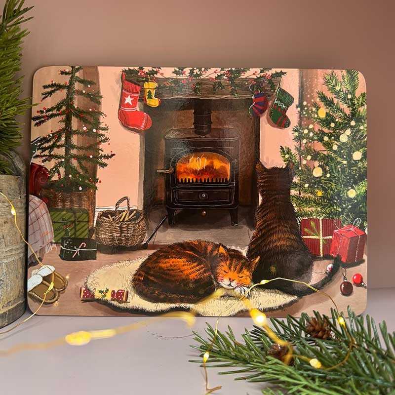 A rectangular decorative placemat featuring a cozy illustrated winter scene with two cats, a fireplace, Christmas stockings, trees, and wrapped gifts