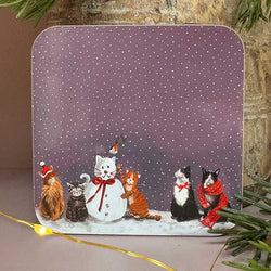 A square decorative coaster with rounded corners featuring a winter scene of five cats around a snowman under a purple snowy sky. 