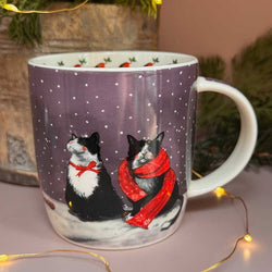 A white ceramic mug with a winter holiday design featuring two black and white cats in snow, one wearing a red bow and the other a red scarf. 