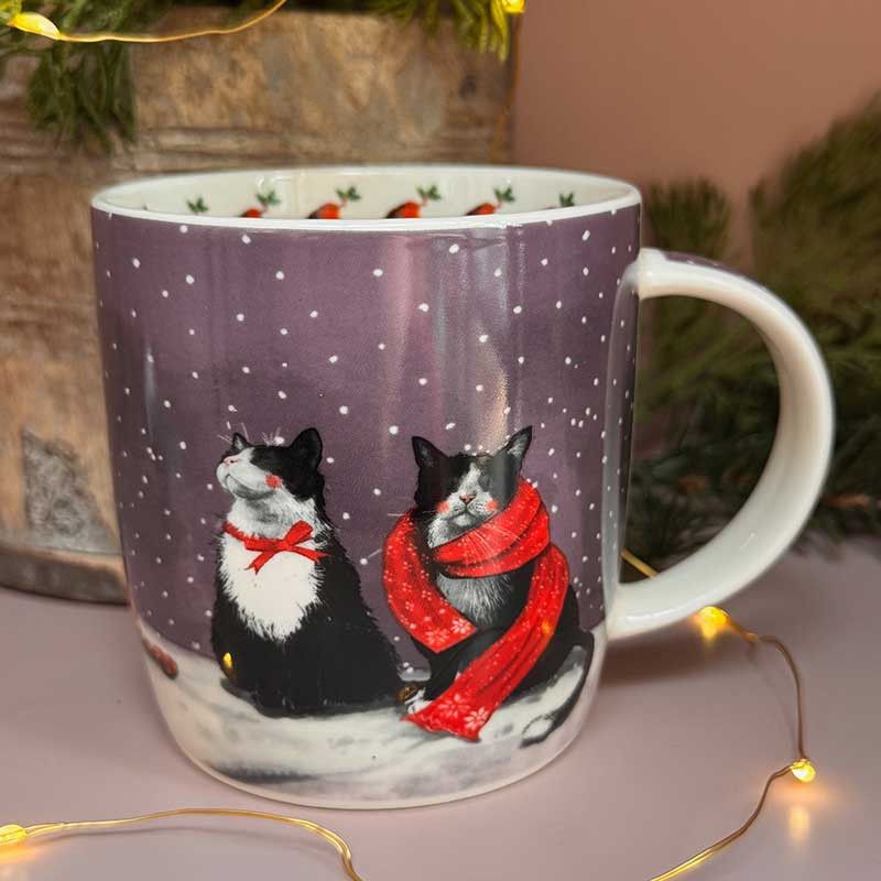 A white ceramic mug with a winter holiday design featuring two black and white cats in snow, one wearing a red bow and the other a red scarf. 