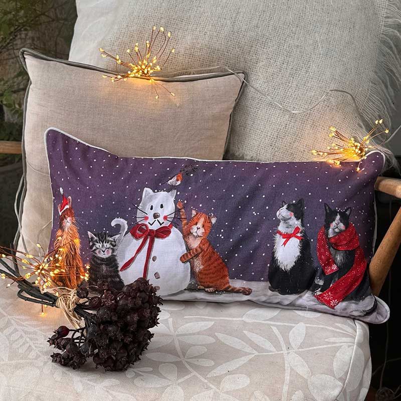 A rectangular throw pillow with a winter holiday design of cats in red scarves and a snowman, styled on a beige armchair with warm fairy lights.