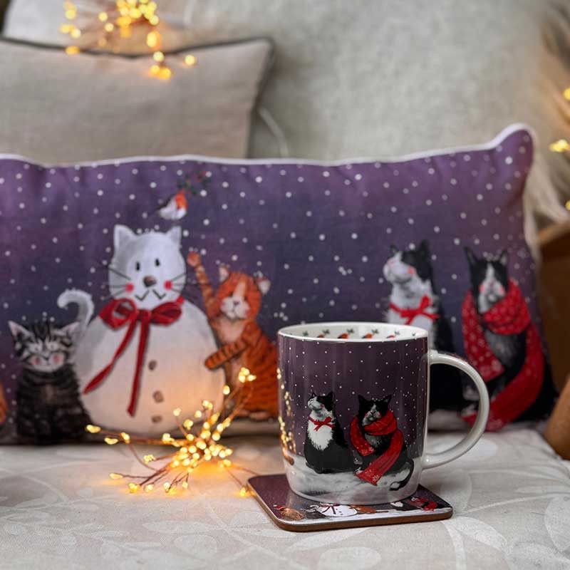 A white ceramic mug with a winter cat print sits on a coaster on a bed, next to a matching purple cat-themed pillow and warm fairy lights.