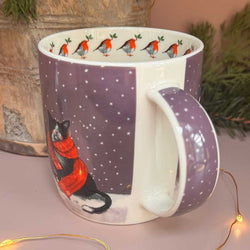 A ceramic mug with a purple winter-themed design featuring a black cat in a red scarf under a starry sky, with a repeating robin pattern on the inner wall