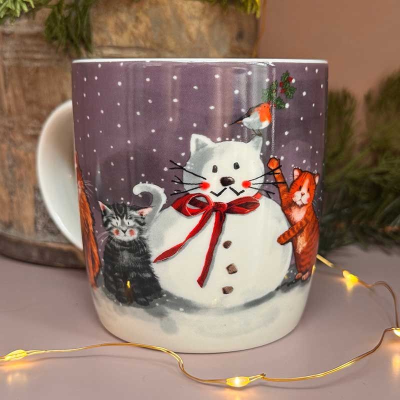 A ceramic mug decorated with a winter holiday scene featuring a snowman, orange tabby cat, black cat, and a small bird on a purple snowy background