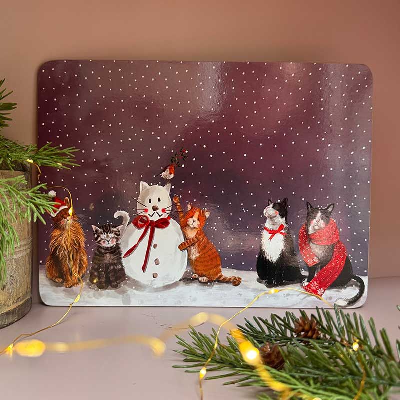 A rectangular placemat featuring five cats dressed in winter clothing, including a snowman, set against a snowy purple background. 