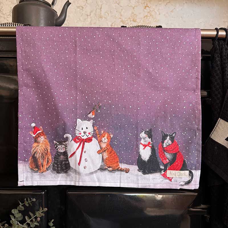 A kitchen towel with a festive winter design, featuring illustrated cats in red scarves and hats, a snowman, and other cats on a snowy ground against a purple sky with white snowflakes.