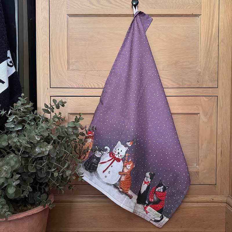 A purple kitchen towel with a white polka-dot pattern and a winter cat illustration, hanging from a black hook on a light wood door next to a potted plant.