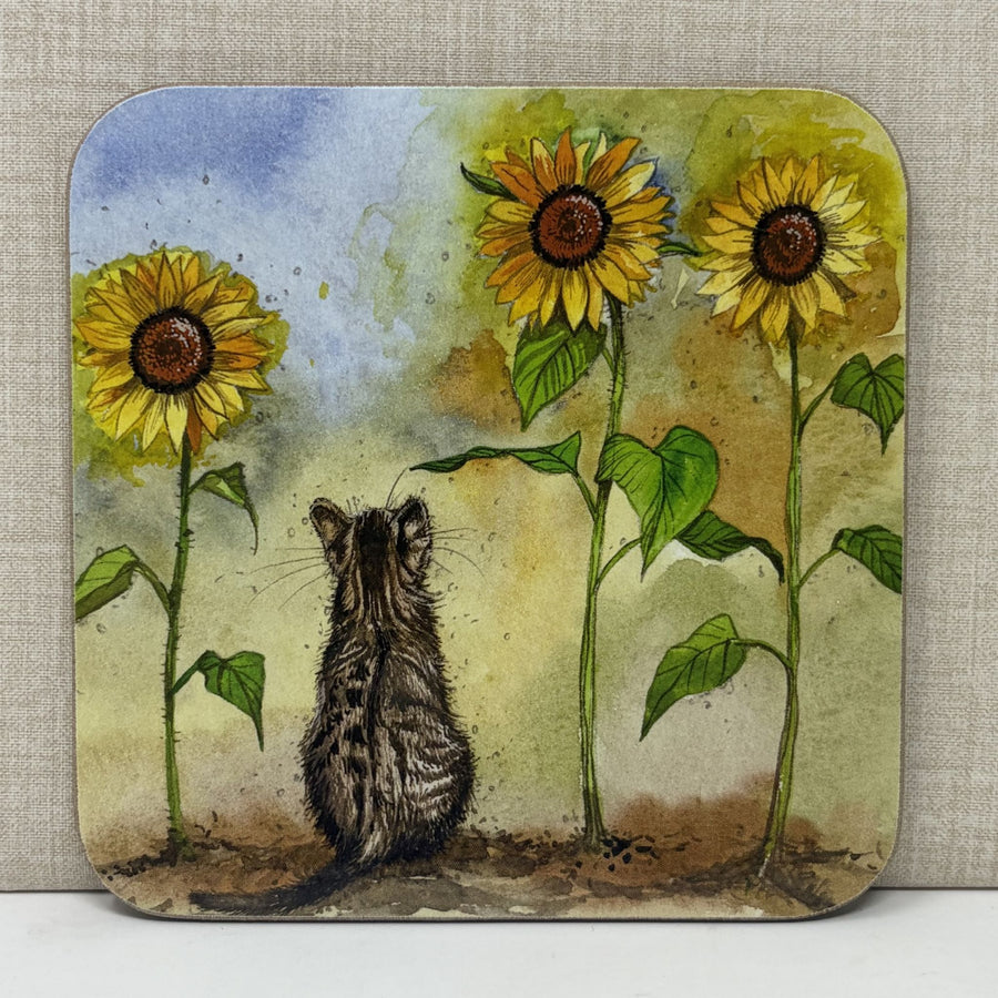 A square coaster with rounded corners featuring a watercolor-style illustration of a small cat sitting in front of three large sunflowers. 