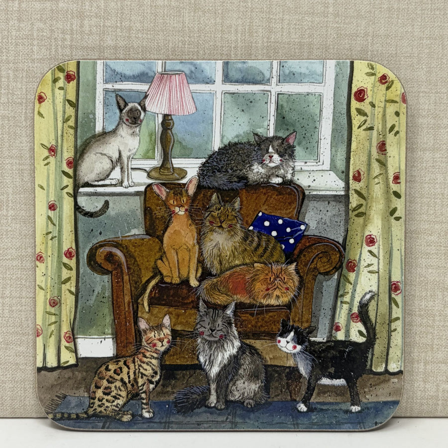 A square coaster with rounded corners featuring a whimsical illustration of six cats lounging in a cosy living room with an armchair, window, lamp, and floral curtains.