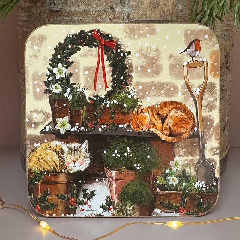 A square decorative coaster featuring a festive Christmas garden scene with a wreath, sleeping cats, flowers, a robin, and a shovel