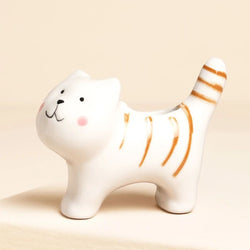 A white ceramic cat-shaped planter with orange stripes, black facial details, and pink cheeks, shown on a warm beige background.