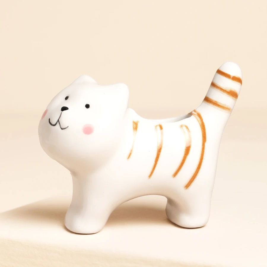 A white ceramic cat-shaped planter with orange stripes, black facial details, and pink cheeks, shown on a warm beige background.